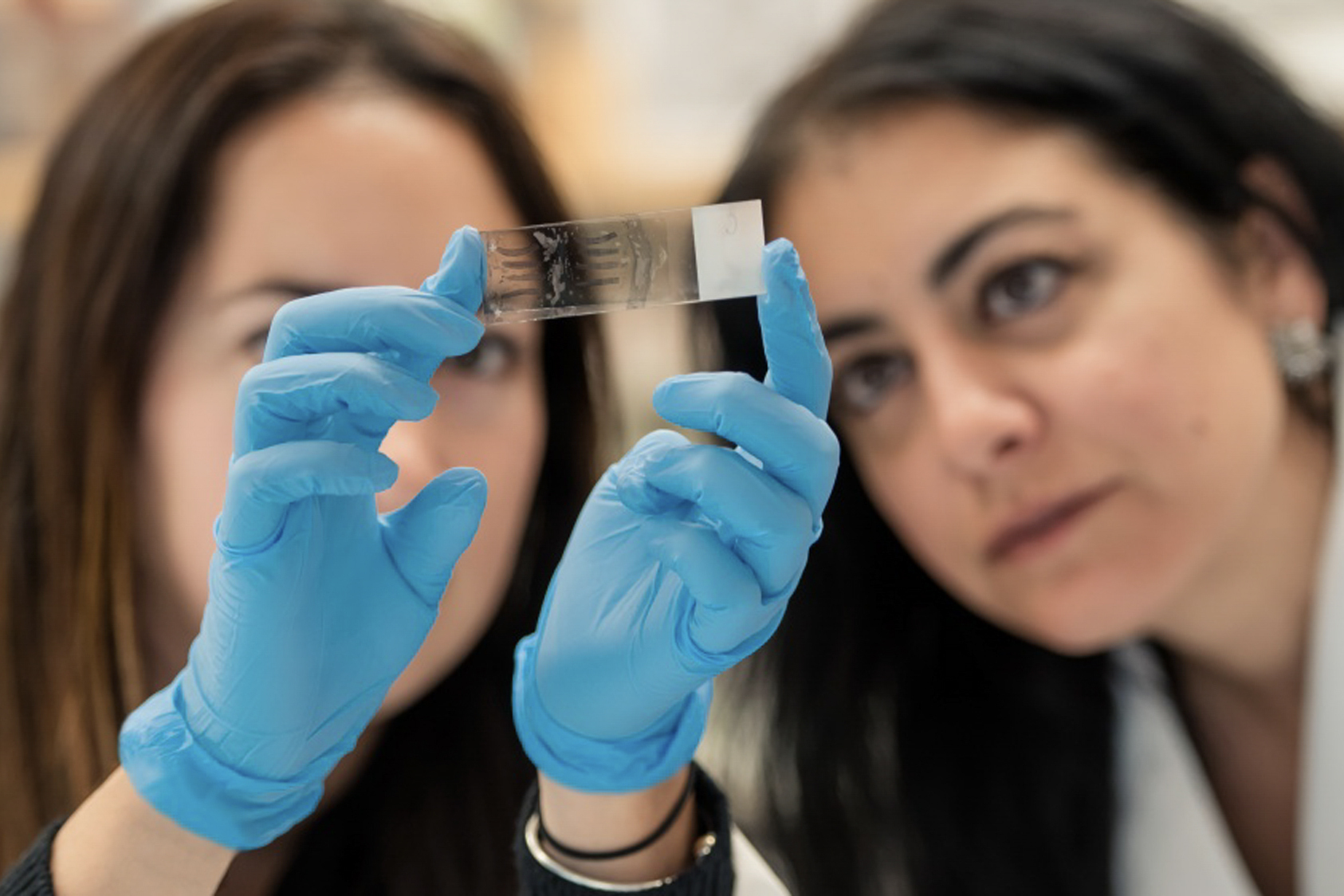 Two people look at a microscope slide being held up by a pair of gloved hands.