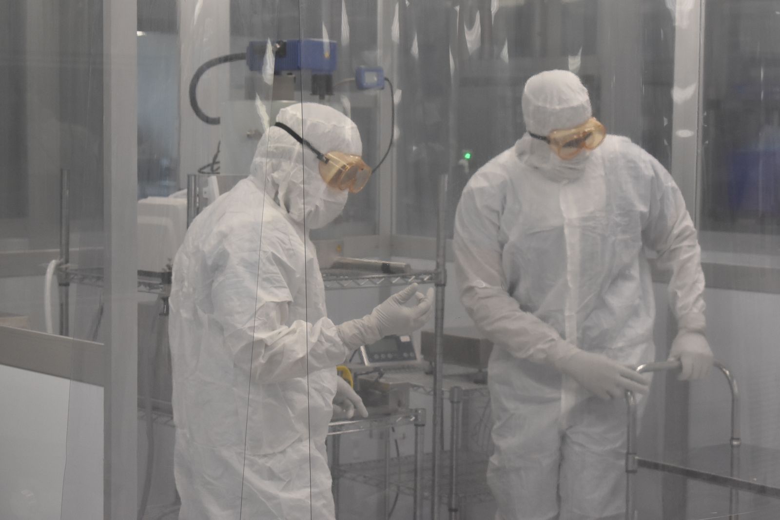 Two workers in white hazmat suits and orange goggles seen through a plastic barrier in a clinical factory setting.