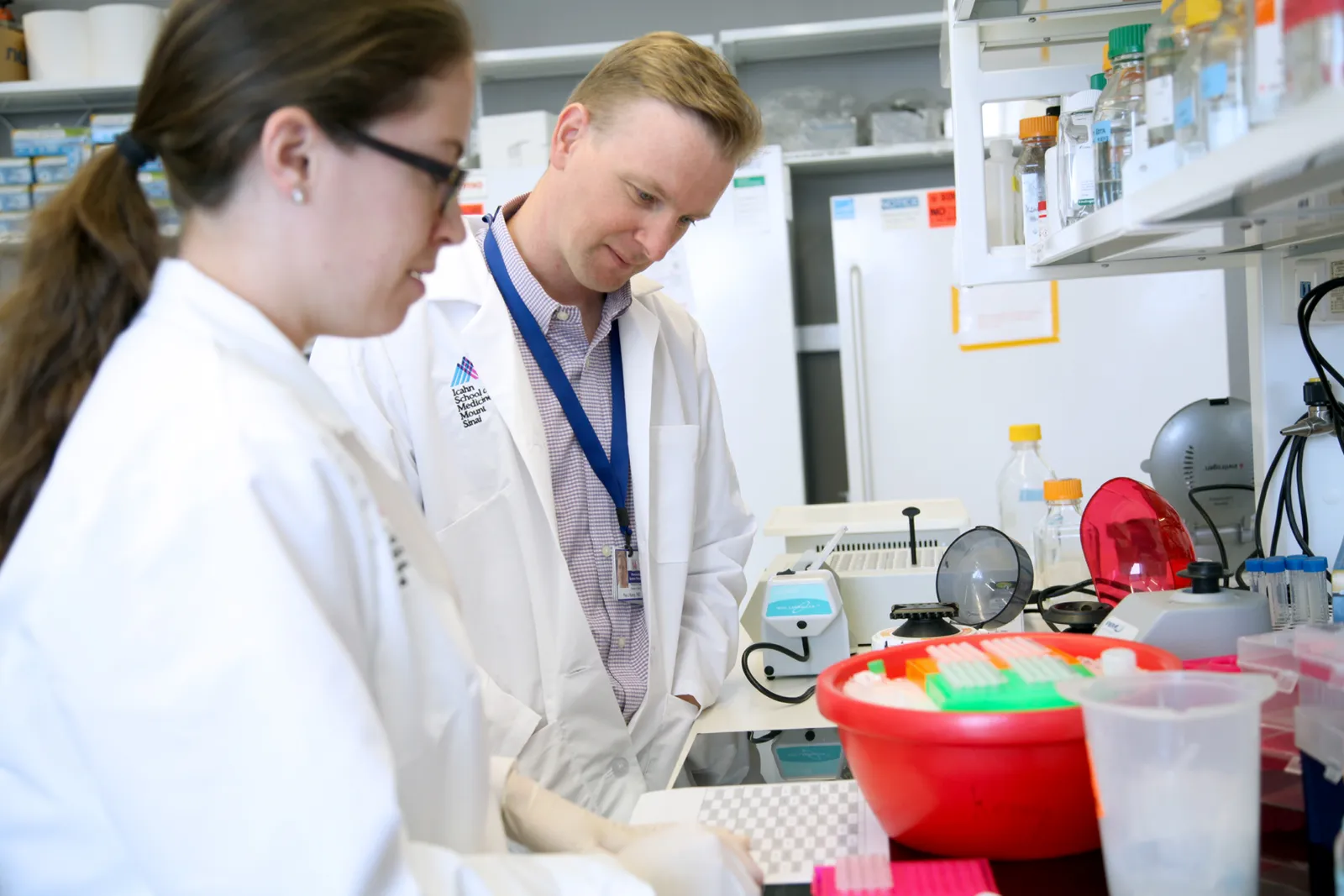 Two people in white coats look at laboratory equipment on a bench.