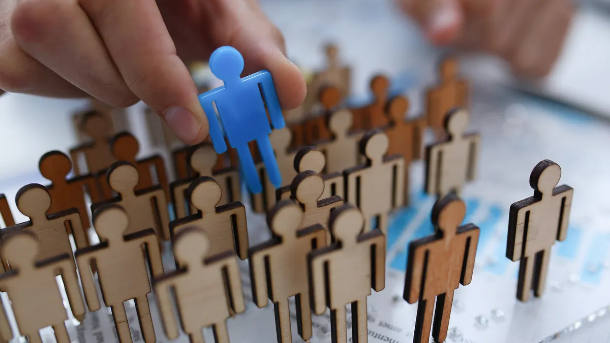 A hand picks a blue figurine out of rows of similar wooden figurines.