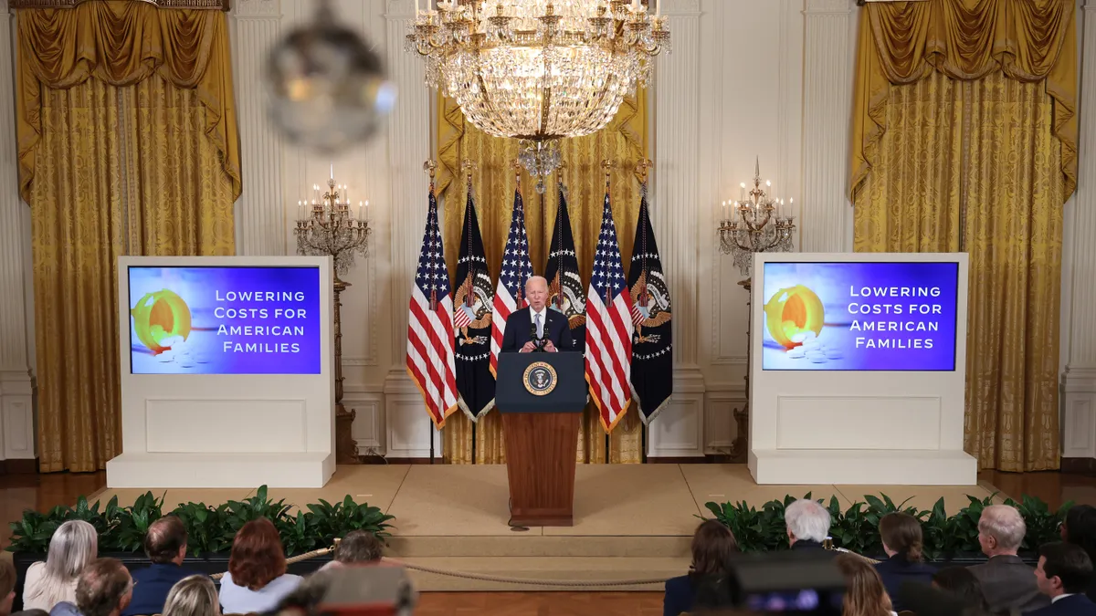 Joe Biden stands at a podium flanked on either sides by screens that read: "lowering costs for American families."