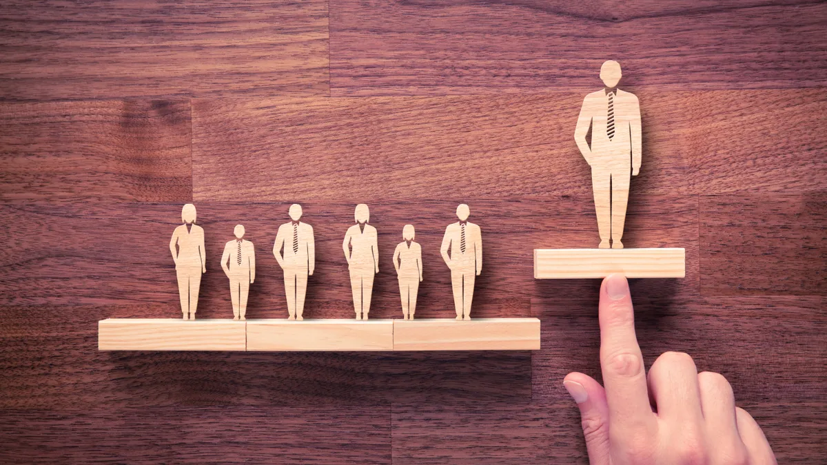 A wooden figurine of a business person sits at a higher spot on a table than a series of six other wooden figurines of business people.